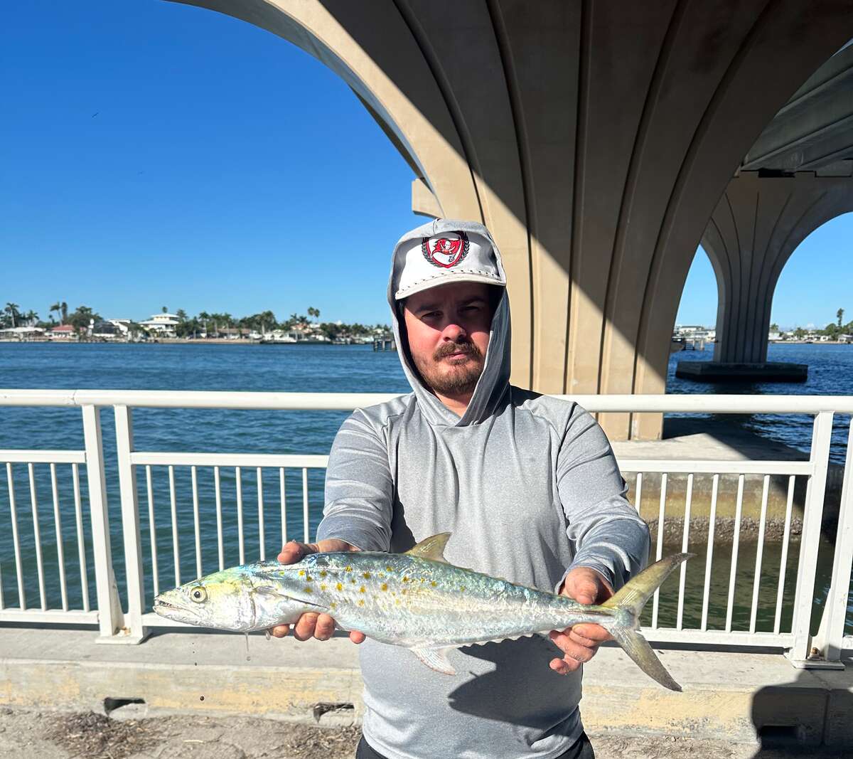 Angler on a bridge pier holding a Spanish mackerel out to the camera, its yellow spots catching the sun