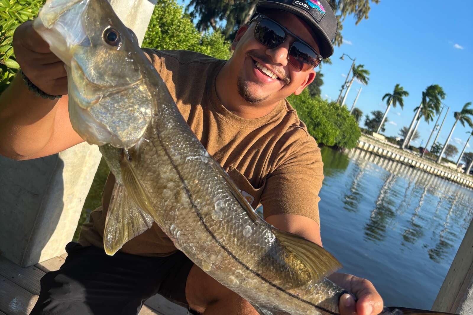 Angler at a Florida dock holding a snook up to the camera, palm trees along the water behind him