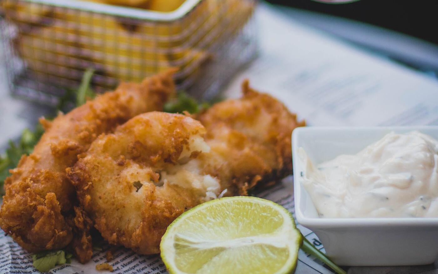 Crispy fried fish pieces with a basket of chips, tartar sauce, and a squeezed lime on newspaper