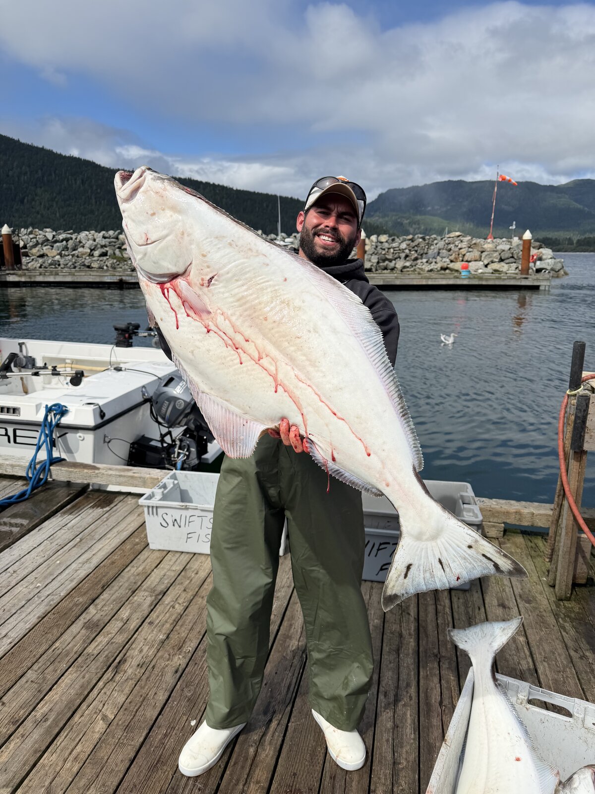 Angler holding a Pacific halibut on a dock with forested hills behind him